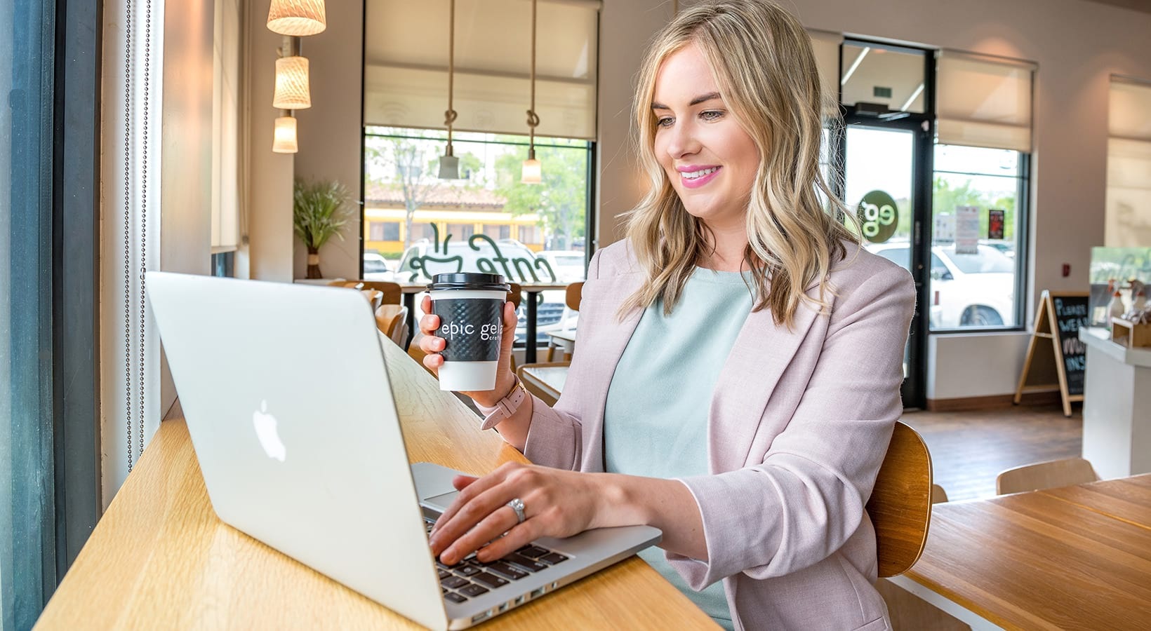 Work From Home  Photograph of someone sitting at their computer at a cafe. They are wearing a pink suit and have a coffee in their hand.